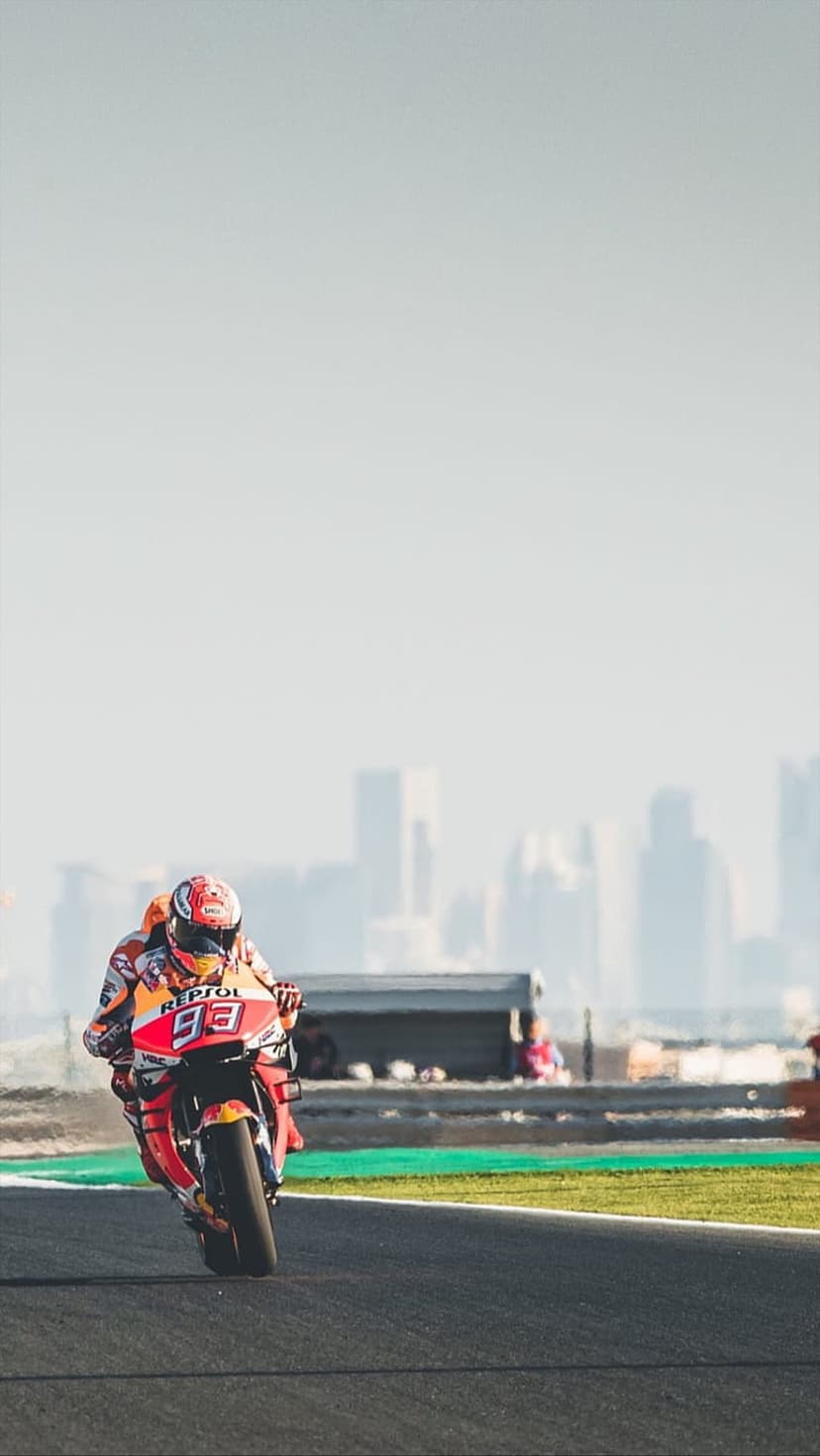 Marc Marquez leans his number 93 Repsol Honda bike into a turn on a racetrack against a distant city skyline.