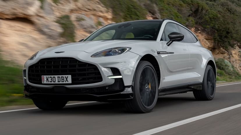 White Aston Martin DBX luxury SUV speeding on a coastal road with motion blur on wheels and cliff background.