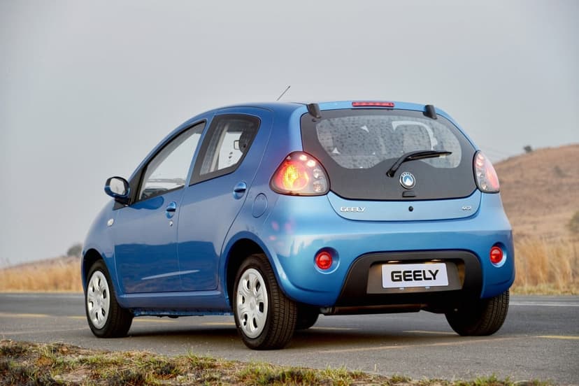 Rear three-quarter view of a blue Geely GC2 city car on a paved road against a soft grassy hillside.