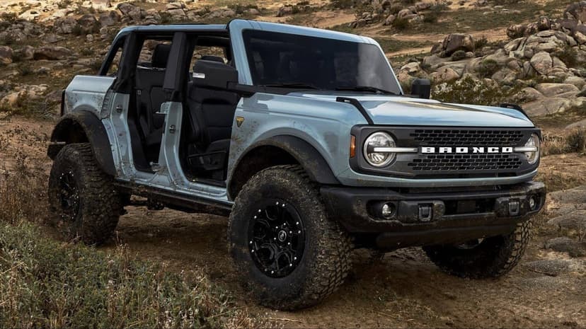 Muddy light blue Ford Bronco with doors removed driving through a rocky desert landscape in natural daylight.