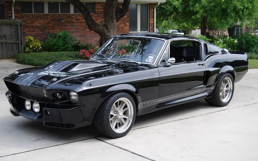 Black 1967 Shelby GT500 Eleanor with grey stripes parked on a driveway in front of a brick house.