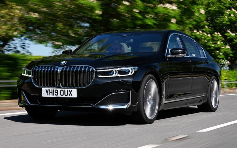 Black BMW 7 Series sedan driving on a road with motion blurred trees and chrome accents under bright daylight.
