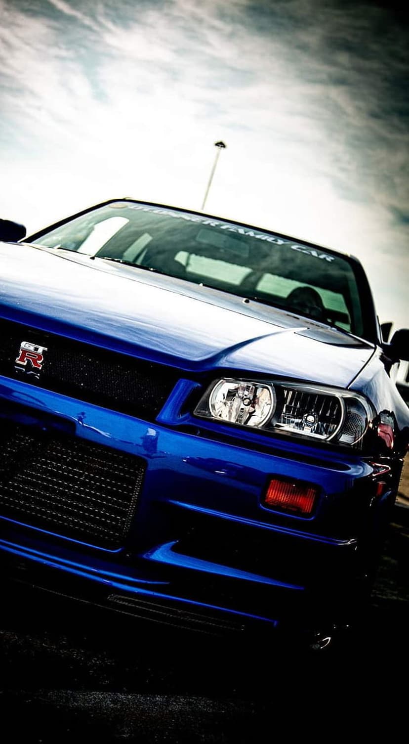 Blue Nissan Skyline R34 GT-R front grille with an R logo and headlights under a dramatic, cloudy sky background.