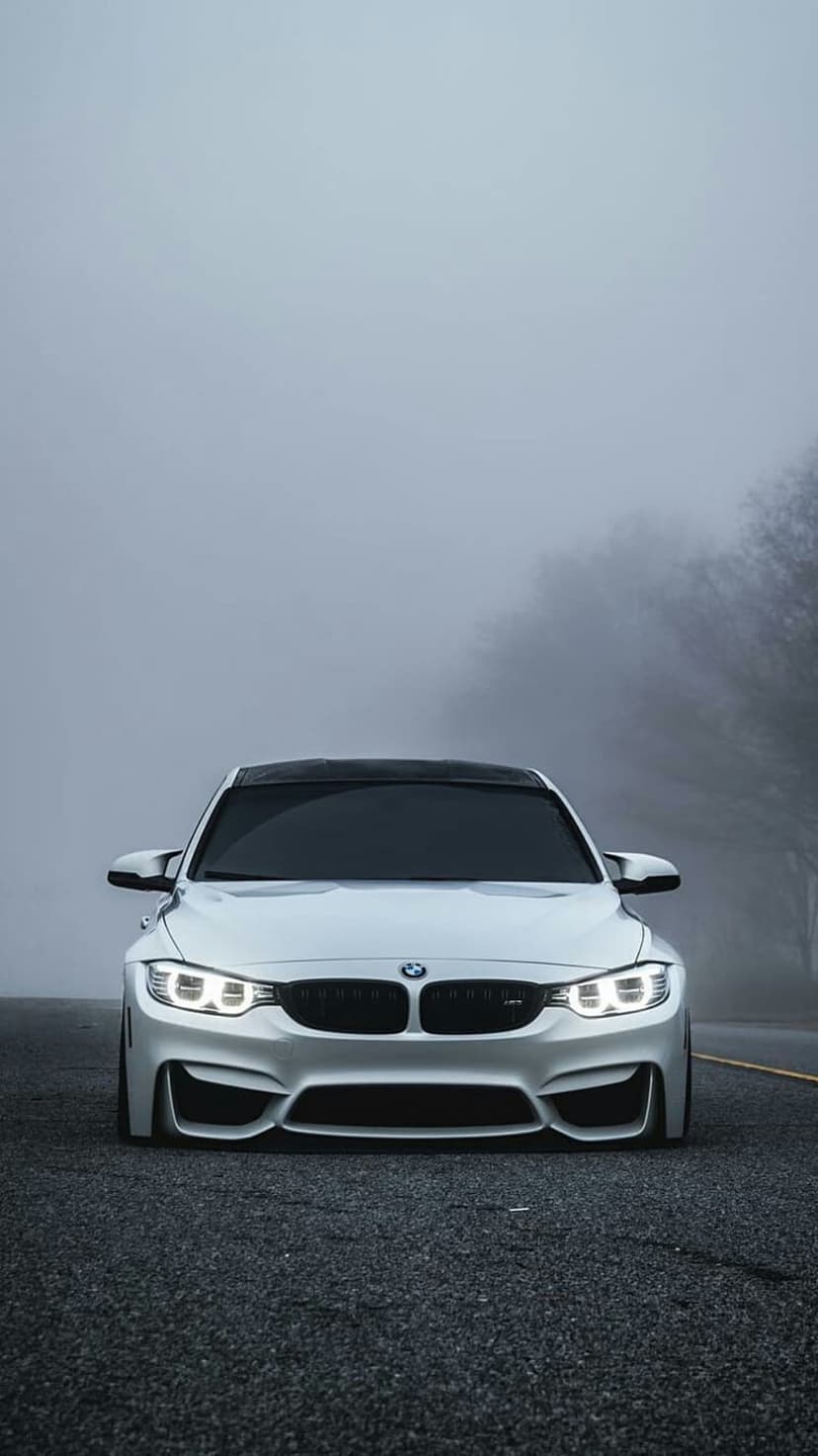 White BMW M4 coupe with lit headlights on a dark road surrounded by heavy fog and blurred trees in the distance.