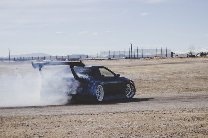 Dark sports car with a large rear wing drifts on a dirt road while emitting thick white smoke near mountains.