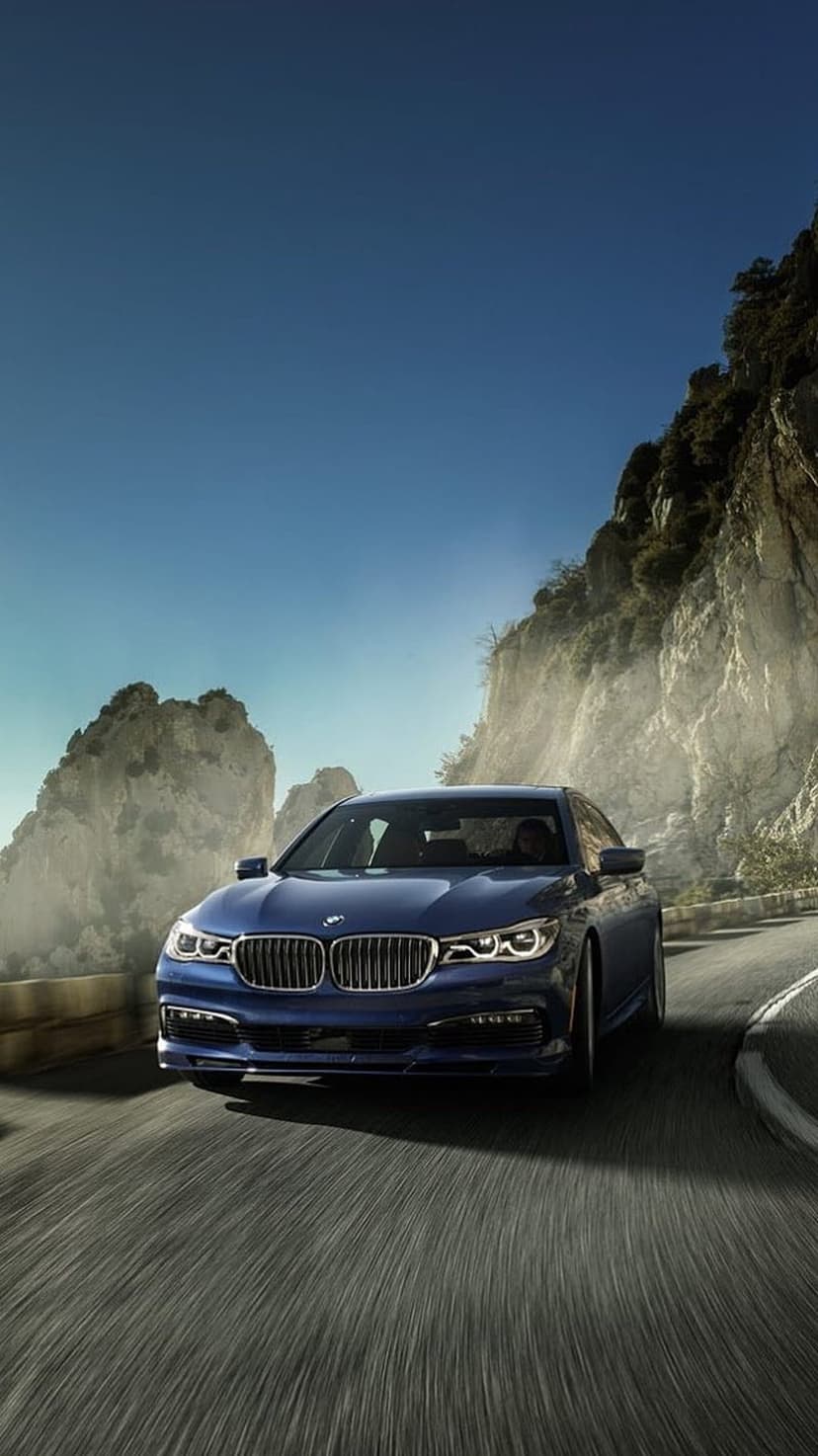 Navy blue luxury sedan driving on a curving mountain road with steep cliffs and a clear blue sky background.