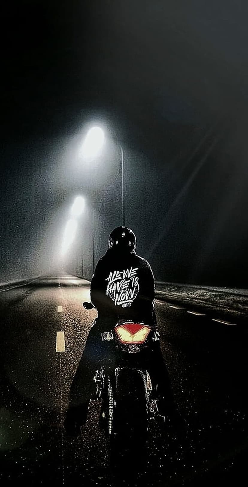 Motorcyclist in a jacket reading All We Have Is Now riding on a wet asphalt road under streetlights at night.