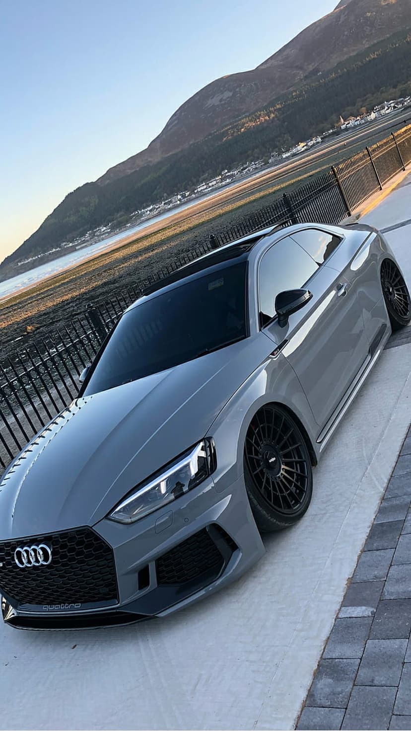Grey Audi RS5 Coupe with custom black wheels parked near a mountain coastline and calm blue sea water.