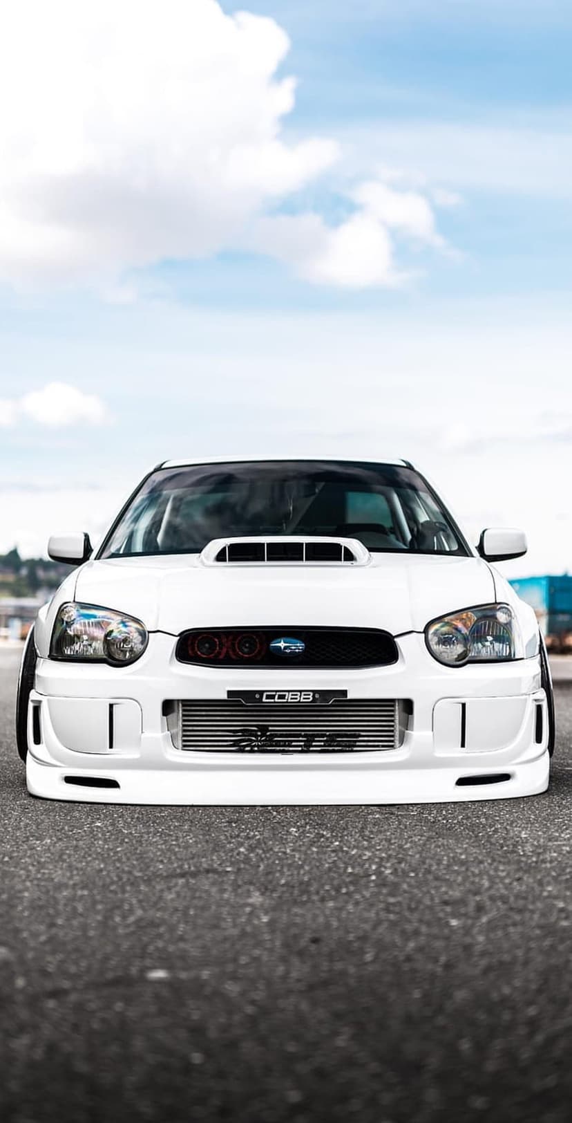 White Subaru WRX STI with a COBB intercooler parked on asphalt under a bright blue sky with white clouds.