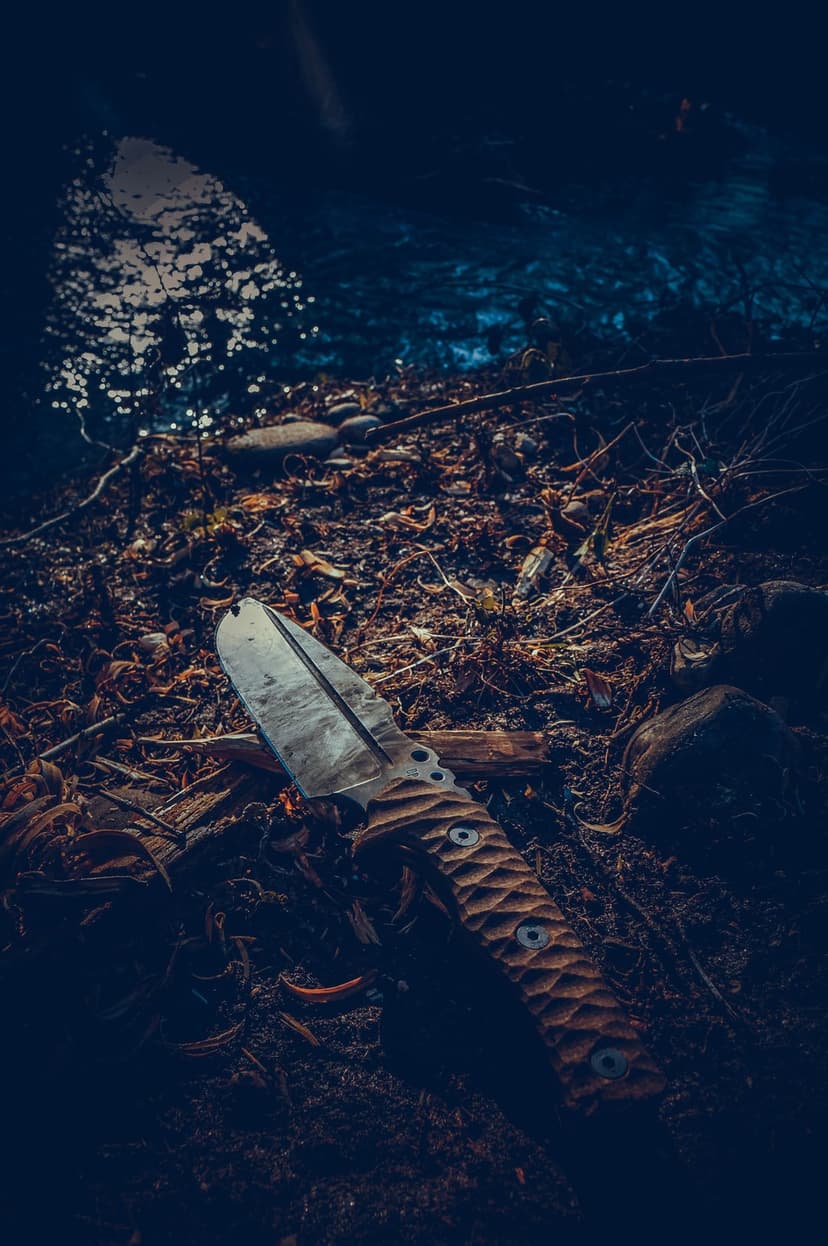 Tactical knife with a textured handle rests on a bed of damp autumn leaves beside a dark flowing forest stream.