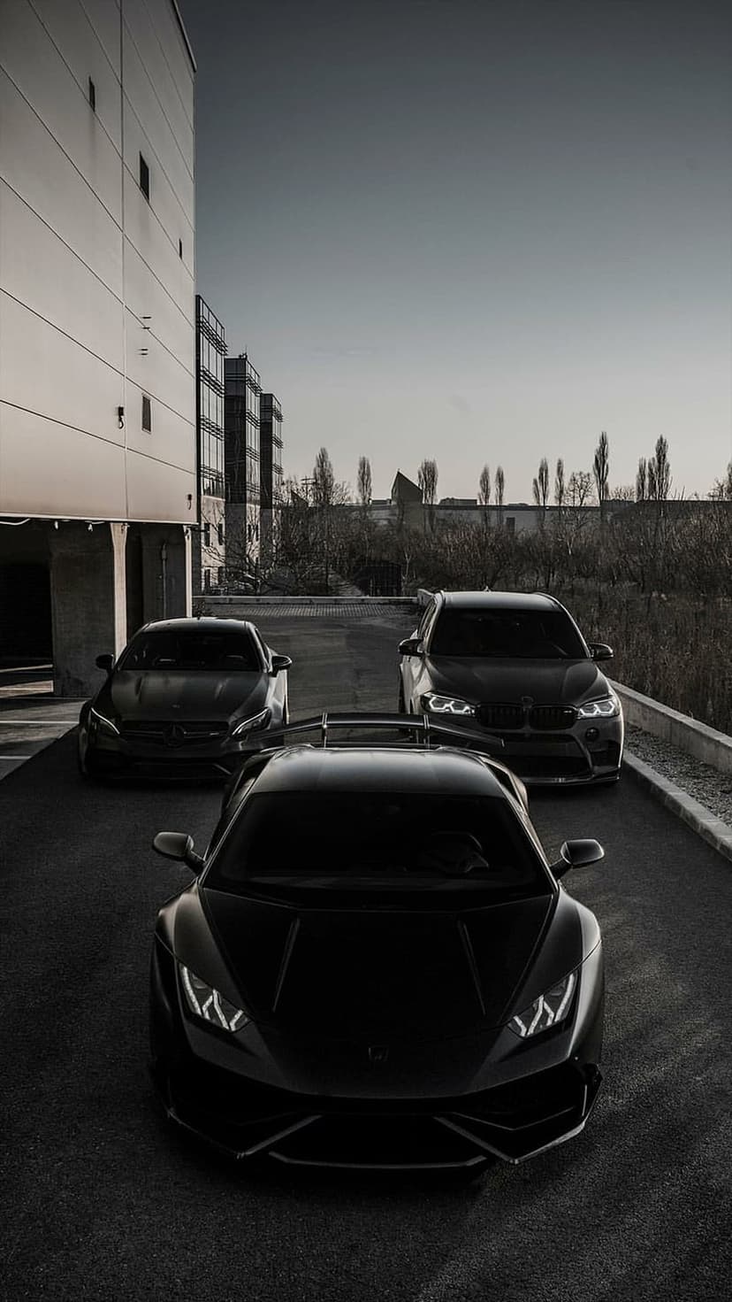 Three black luxury cars including a supercar and SUV parked in a row near an industrial building at twilight.