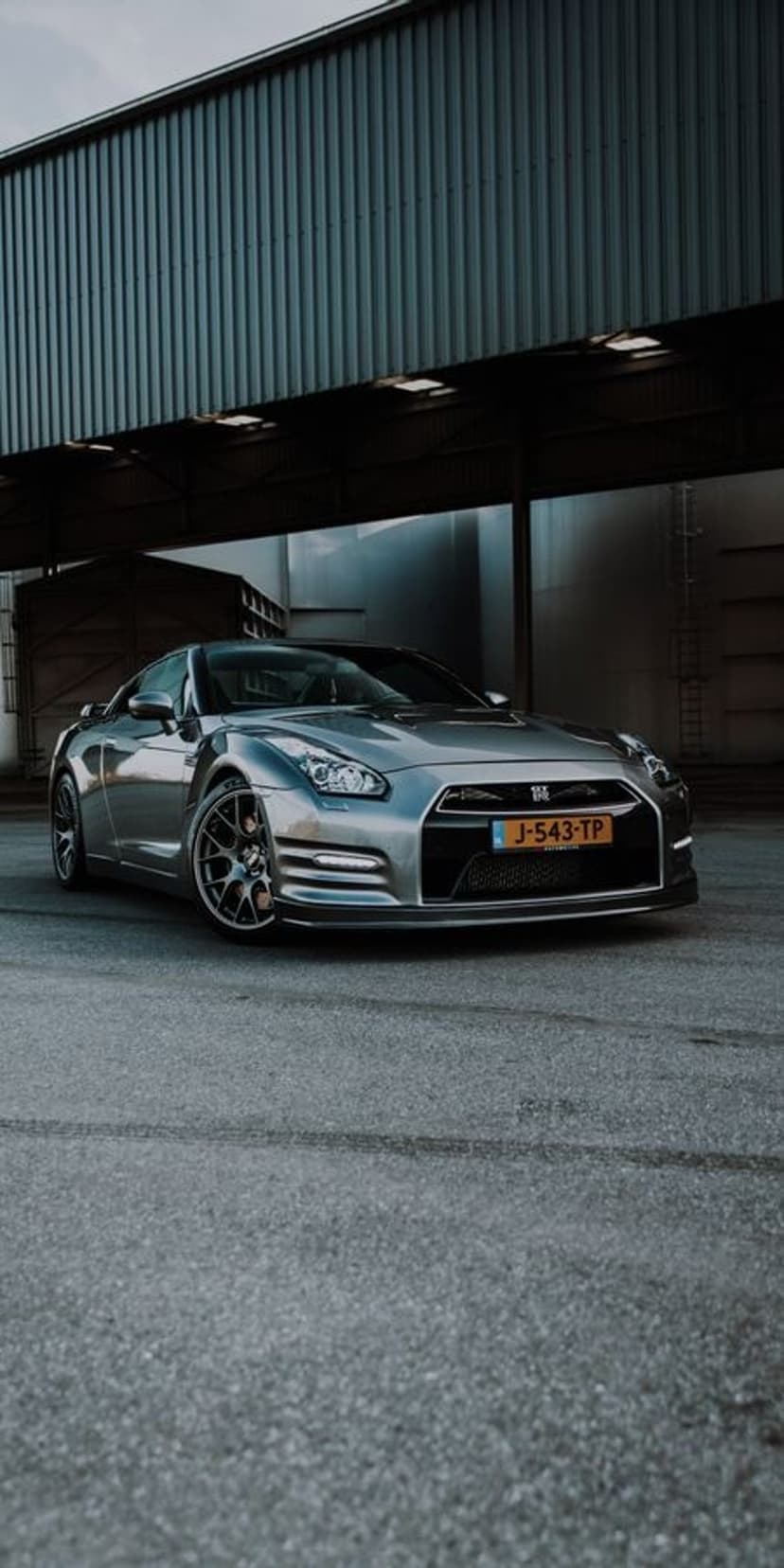 Silver Nissan GT-R R35 parked on asphalt under a corrugated metal building shadow at a low camera angle.
