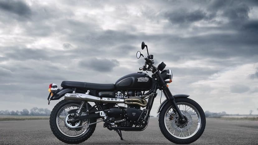 Profile of a black Triumph Scrambler motorcycle parked on open asphalt under a moody and dramatic overcast sky.