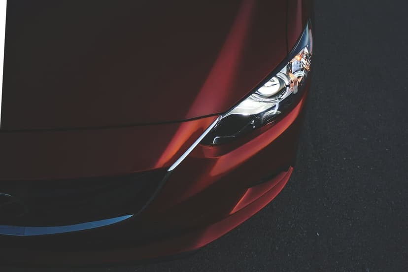 Red sports car headlight and sculpted hood featuring sharp sun shadows and metallic chrome accents on asphalt.