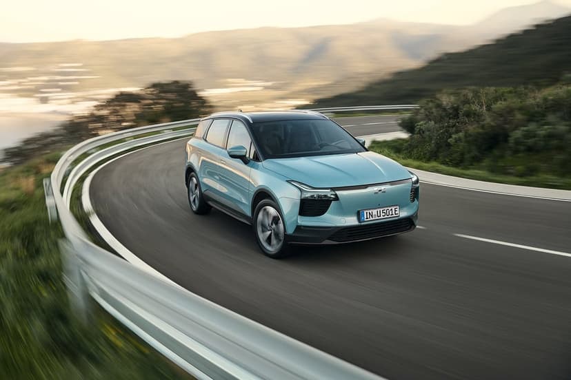 Light blue electric SUV driving on a curved road with motion blur and green mountain scenery background.