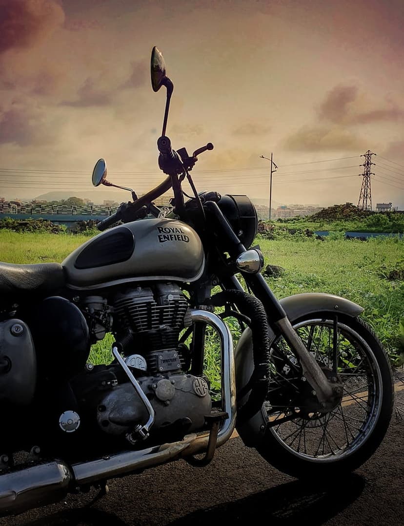 Royal Enfield motorcycle with chrome engine details parked on a roadside under a dark, cloudy sky by green fields.
