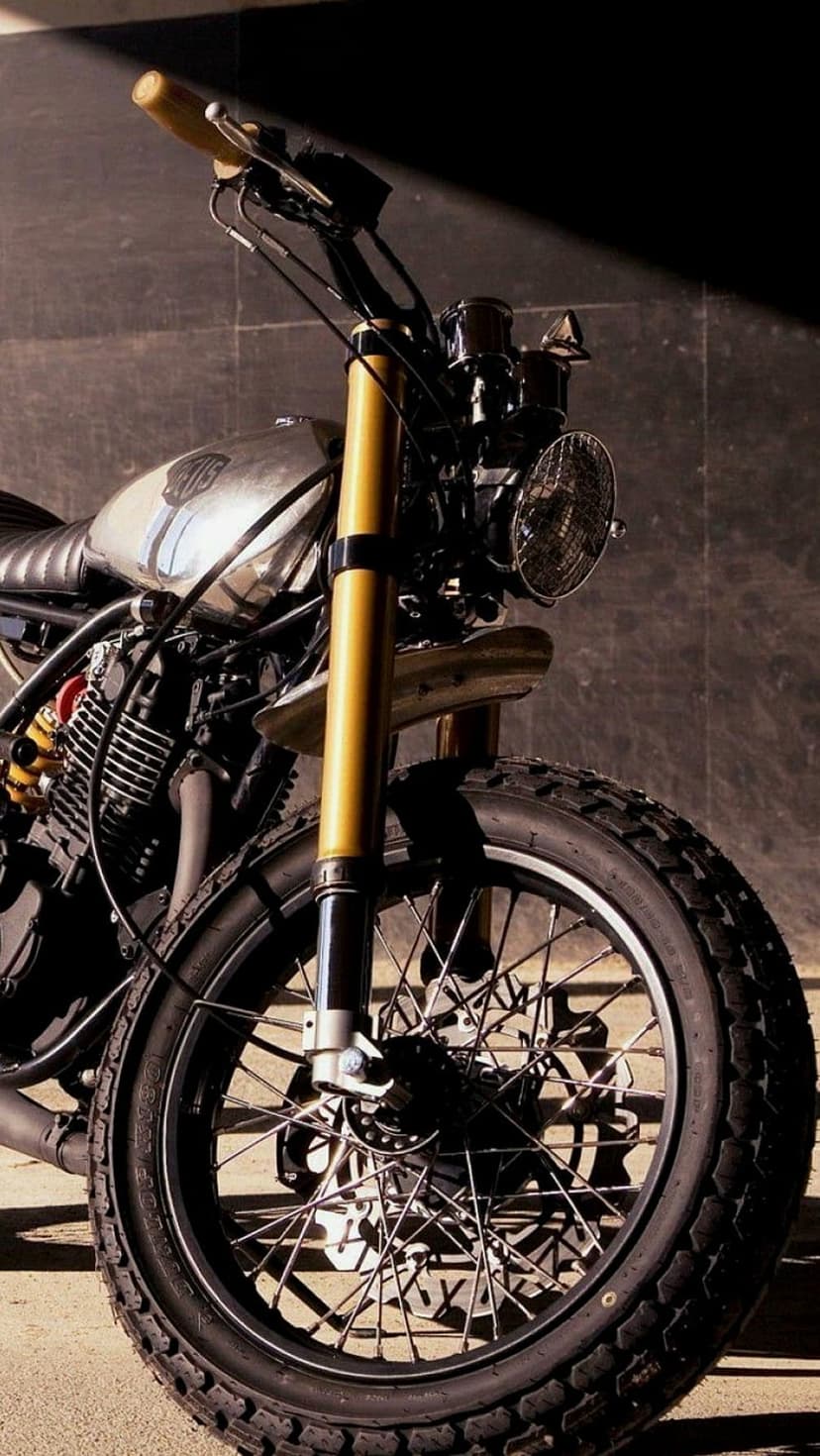 Custom motorcycle front wheel with golden forks, a chrome fuel tank, and knobby tires on a wire-spoke rim.
