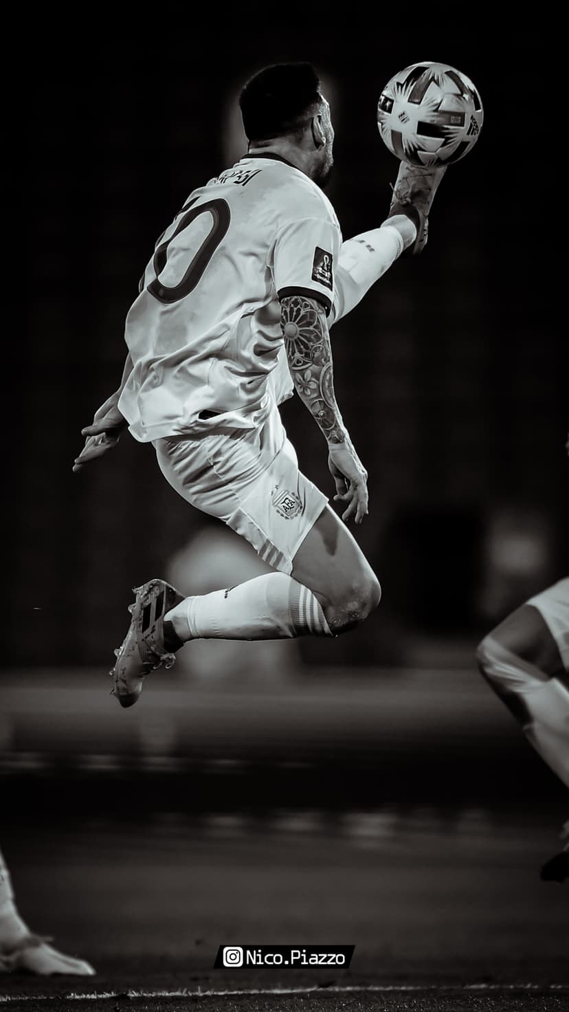 Lionel Messi leaps mid-air to control a football, displaying arm tattoos in a high-contrast monochrome scene.