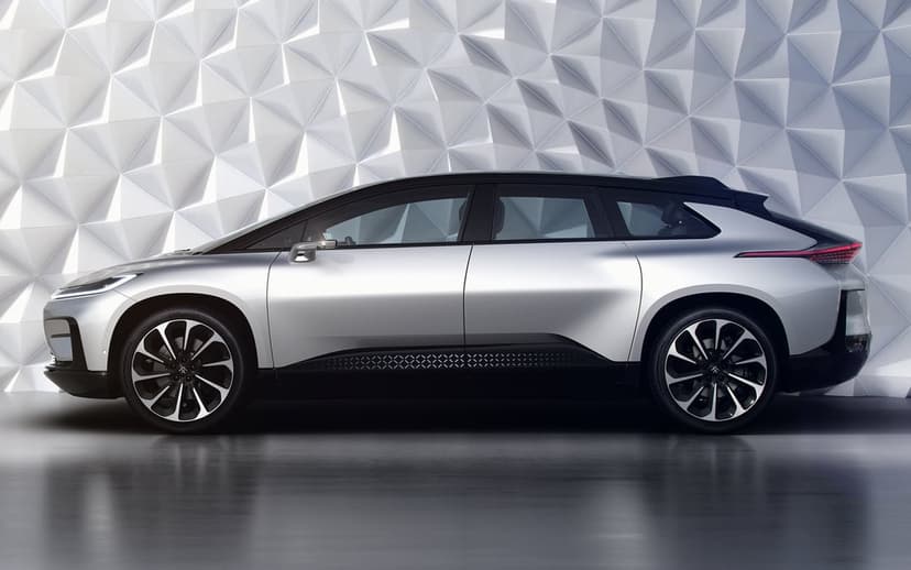 Silver electric SUV profile view parked in front of a white 3D faceted wall with soft studio lighting.