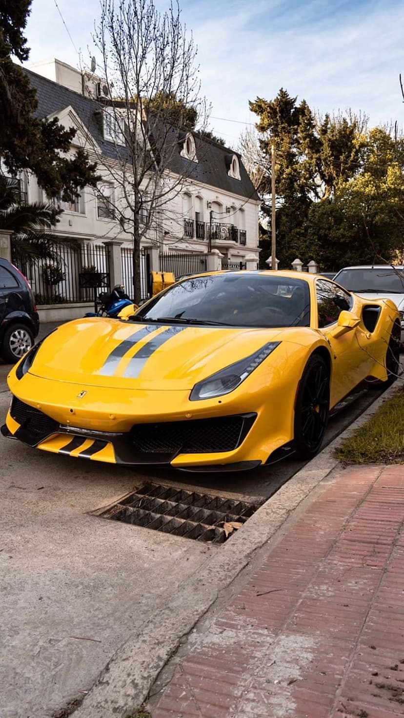 Yellow Ferrari with silver racing stripes parked on a street in front of a minimalist modern white house.