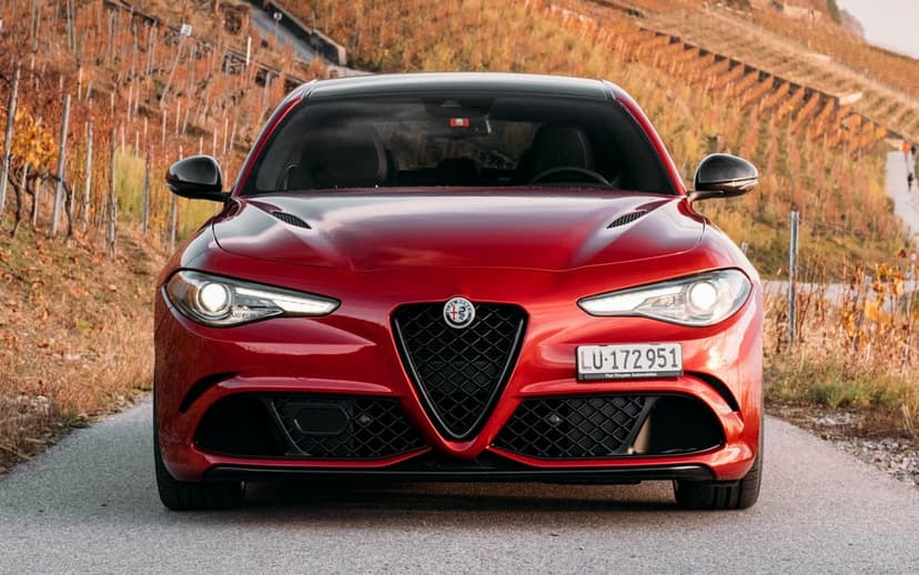 Front view of a red Alfa Romeo sports sedan on a paved road with autumn vineyards and golden hour light behind.