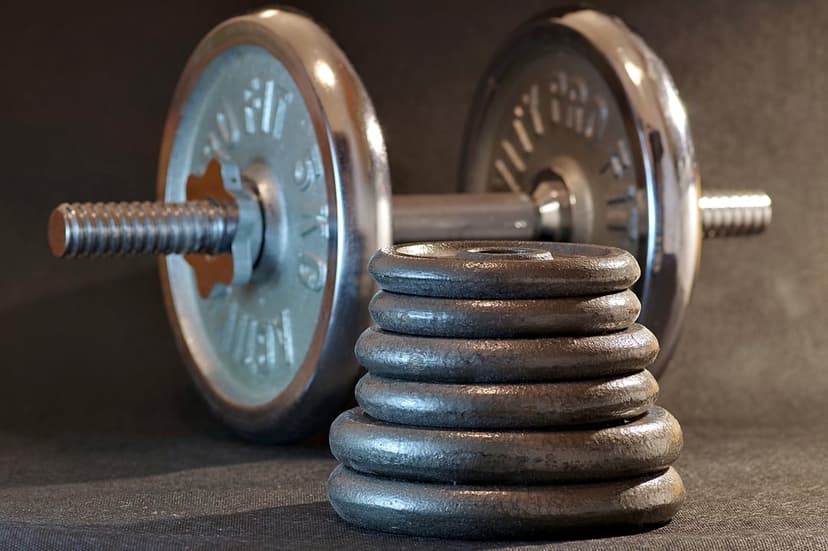 Chrome adjustable dumbbell with a threaded bar sits in front of stacked black cast iron weight plates in a gym.