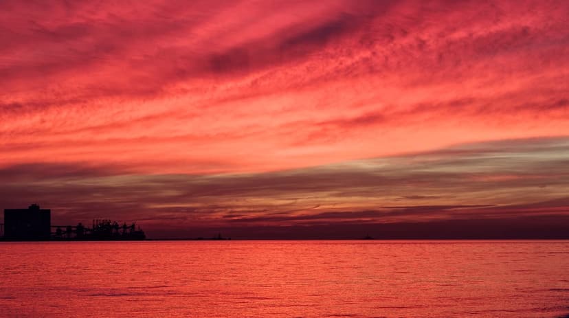 Deep red sunset sky reflects on calm ocean waves behind a dark, distant city skyline silhouette at dusk.