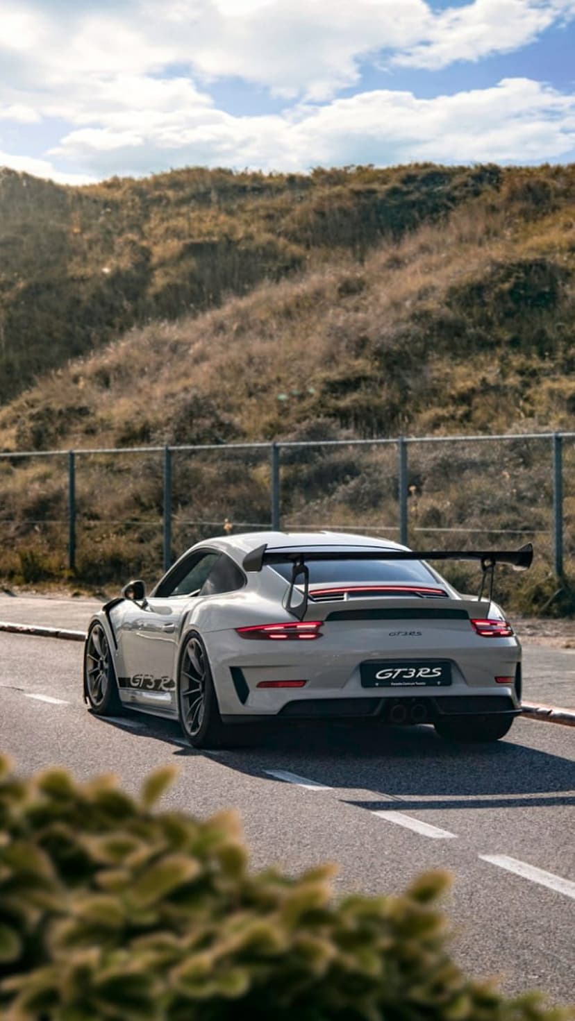 Porsche GT3 RS with a large rear wing parked on a coastal road during golden hour with grass and a fence nearby.