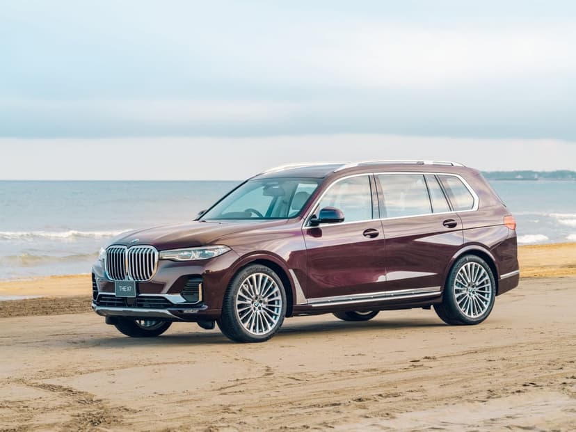 Burgundy BMW X7 SUV on a sandy beach with ocean waves and a soft cloudy sky in the distance.