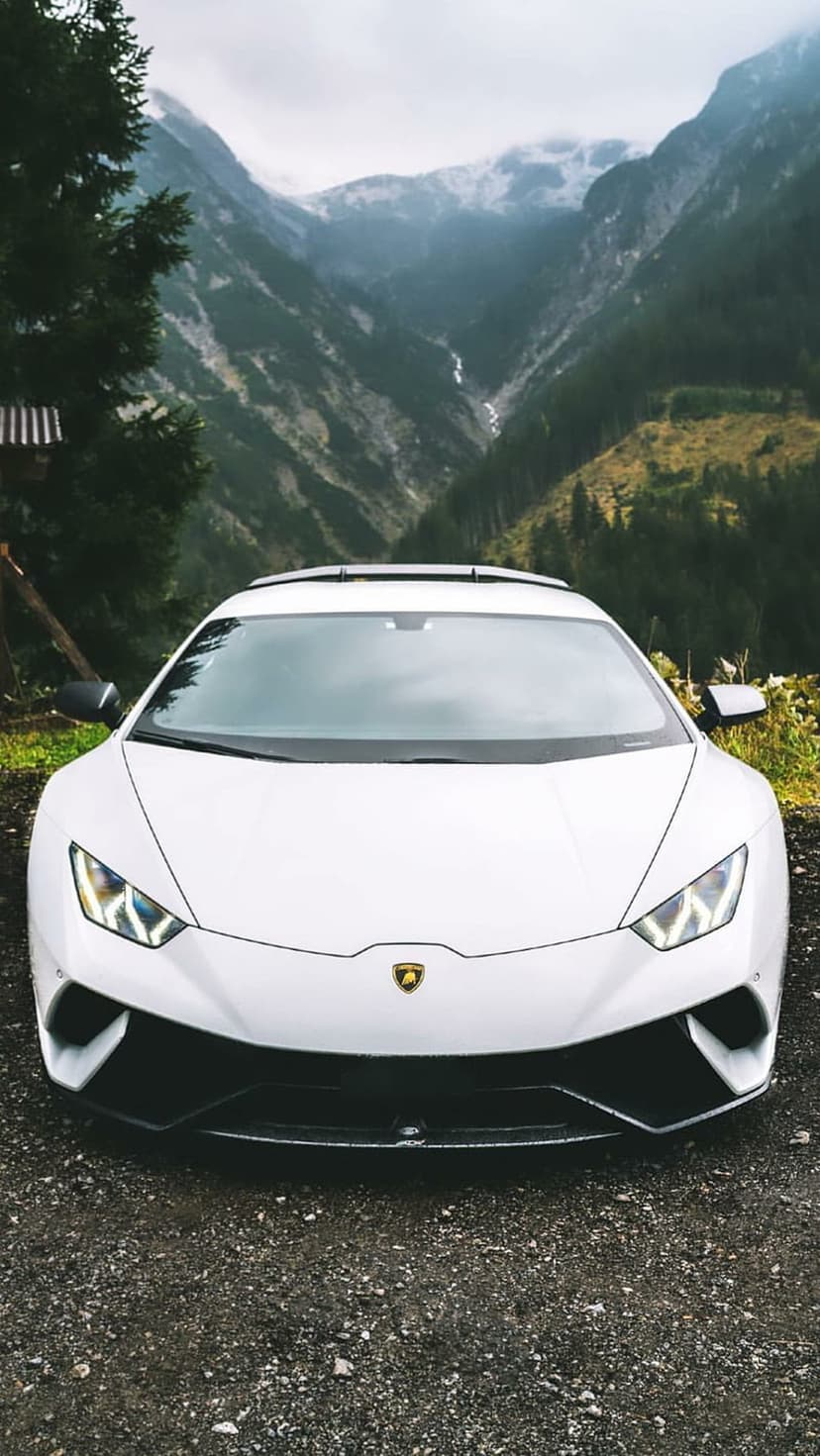 White Lamborghini Huracan on a gravel road with mist-covered mountains and evergreen forests in the background.