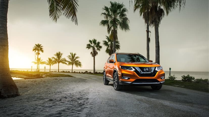 Orange SUV parked on a sandy beach road lined with palm trees under a warm golden sunset.