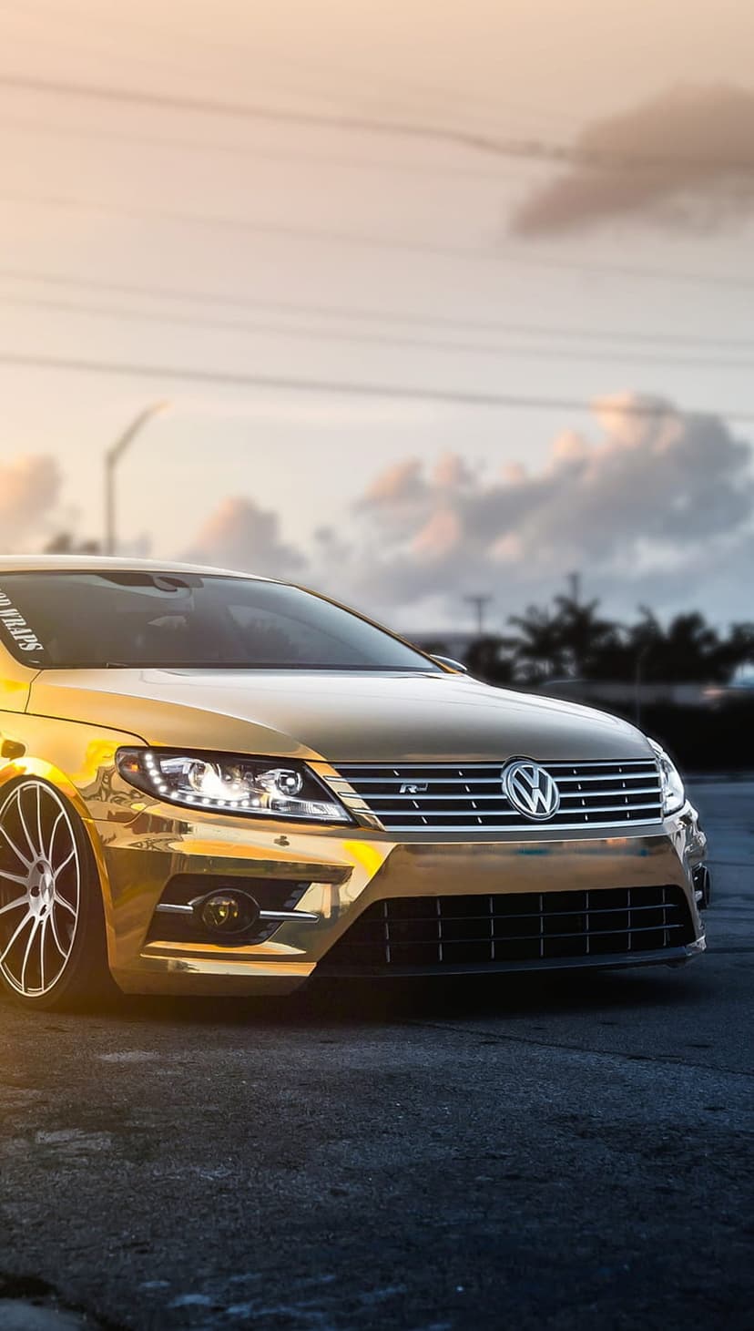 Gold Volkswagen CC with custom wheels and a lowered stance parked against a vibrant orange sunset background.