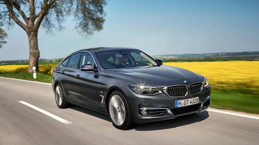 Grey BMW Gran Turismo driving on a rural road beside a yellow flower field under a clear blue sky.