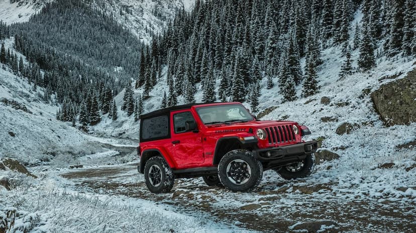 Red Jeep Wrangler Rubicon parked on a rocky snowy mountain trail with pine trees and hills in the background.