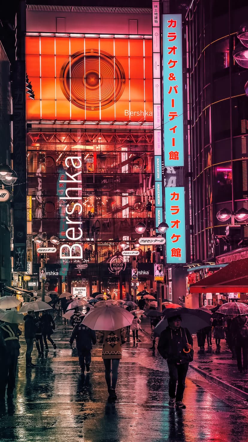 Pedestrians with umbrellas cross a wet Tokyo street illuminated by red and blue neon signs at night.