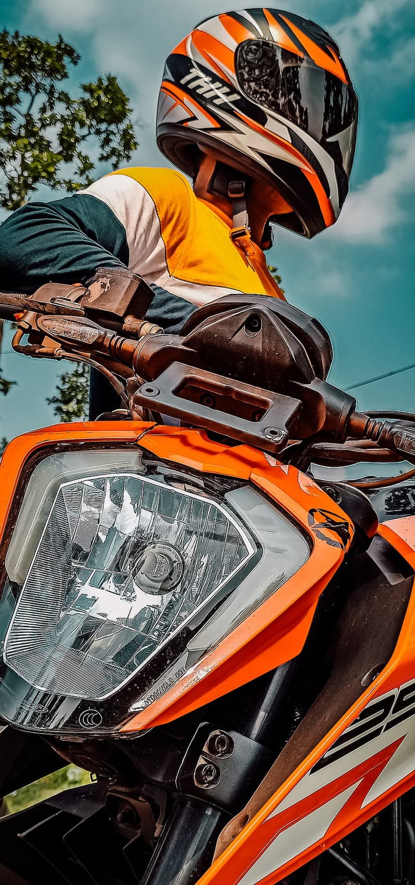 Motorcyclist in an orange helmet sits on a matching sportbike with a cloudy sky and green trees in the background.