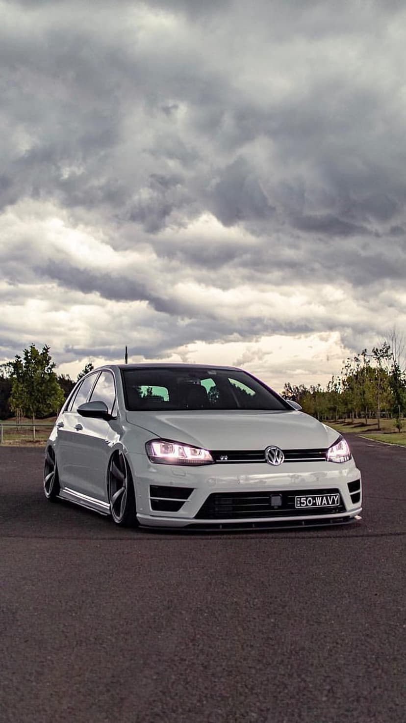 White Volkswagen Golf R with dark rims and lit headlights on asphalt under a dramatic grey cloudy sky.