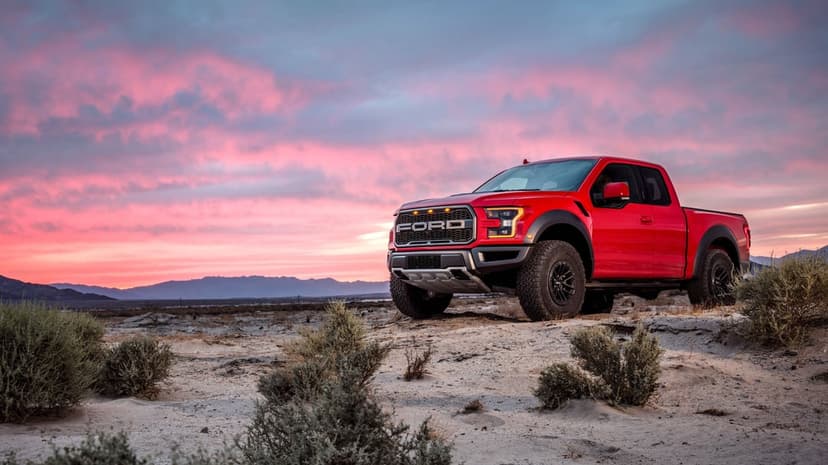 Red off-road truck parked on sandy desert terrain under a vibrant pink and purple sunset sky with mountains.