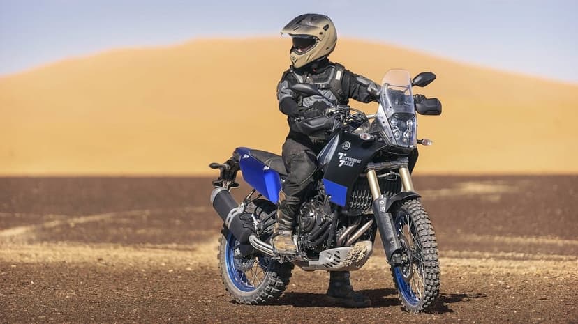 Yamaha Tenere 700 rider in gear on a rocky desert floor with a massive golden sand dune in the background.