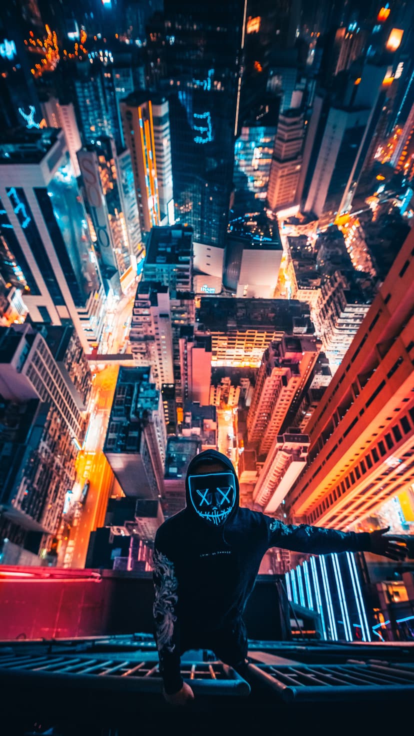 Masked person with glowing XX eyes kneels on a rooftop edge overlooking a vast, illuminated night cityscape.