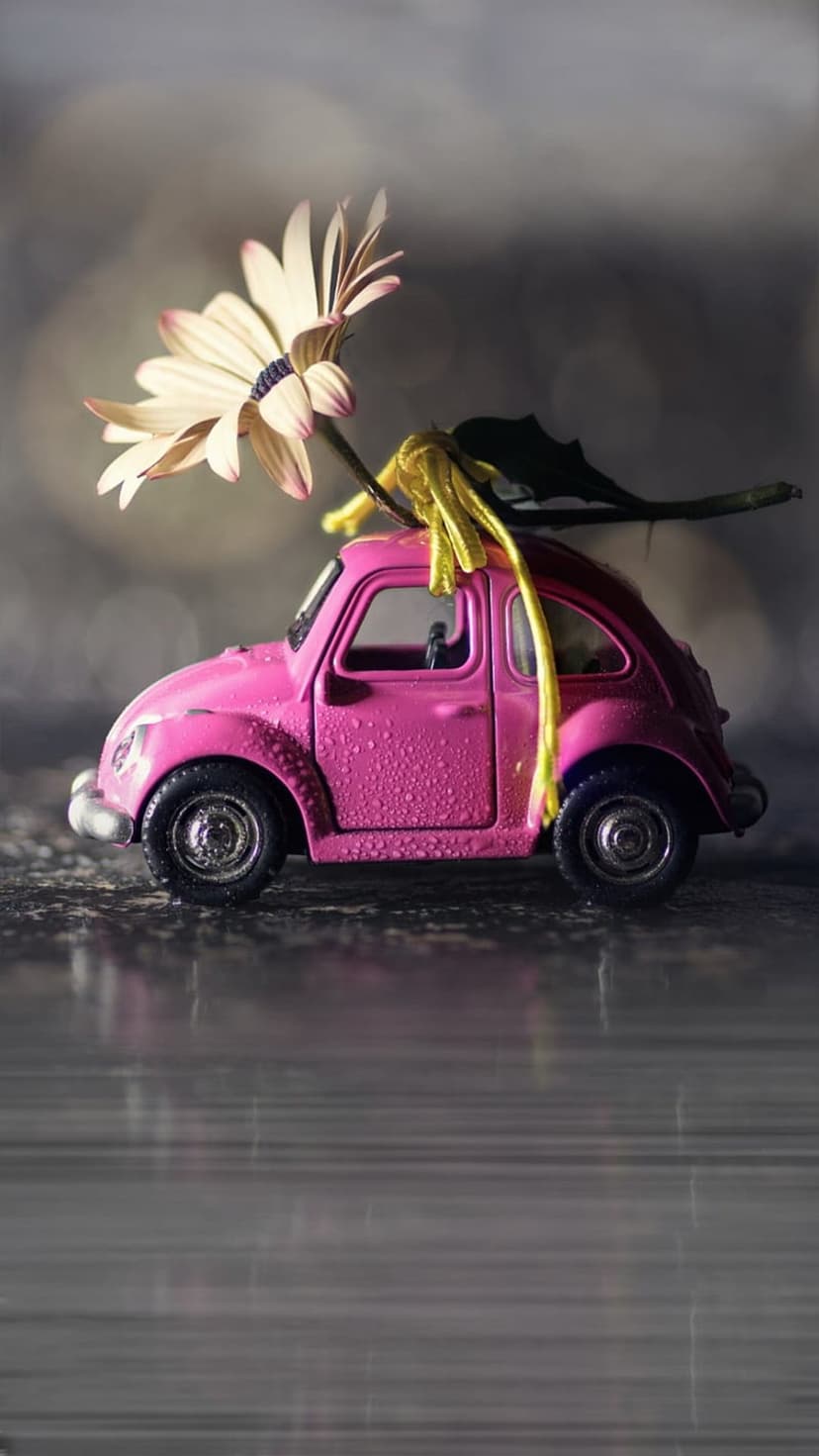 Pink toy Beetle car with rain droplets and a flower on its roof sits before a blurred bokeh light background.