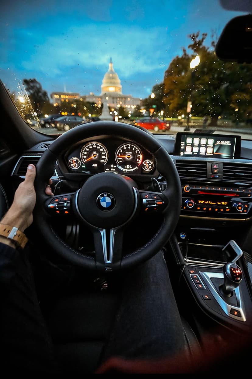 BMW steering wheel and dashboard interior facing the glowing U.S. Capitol Building through a rainy windshield.