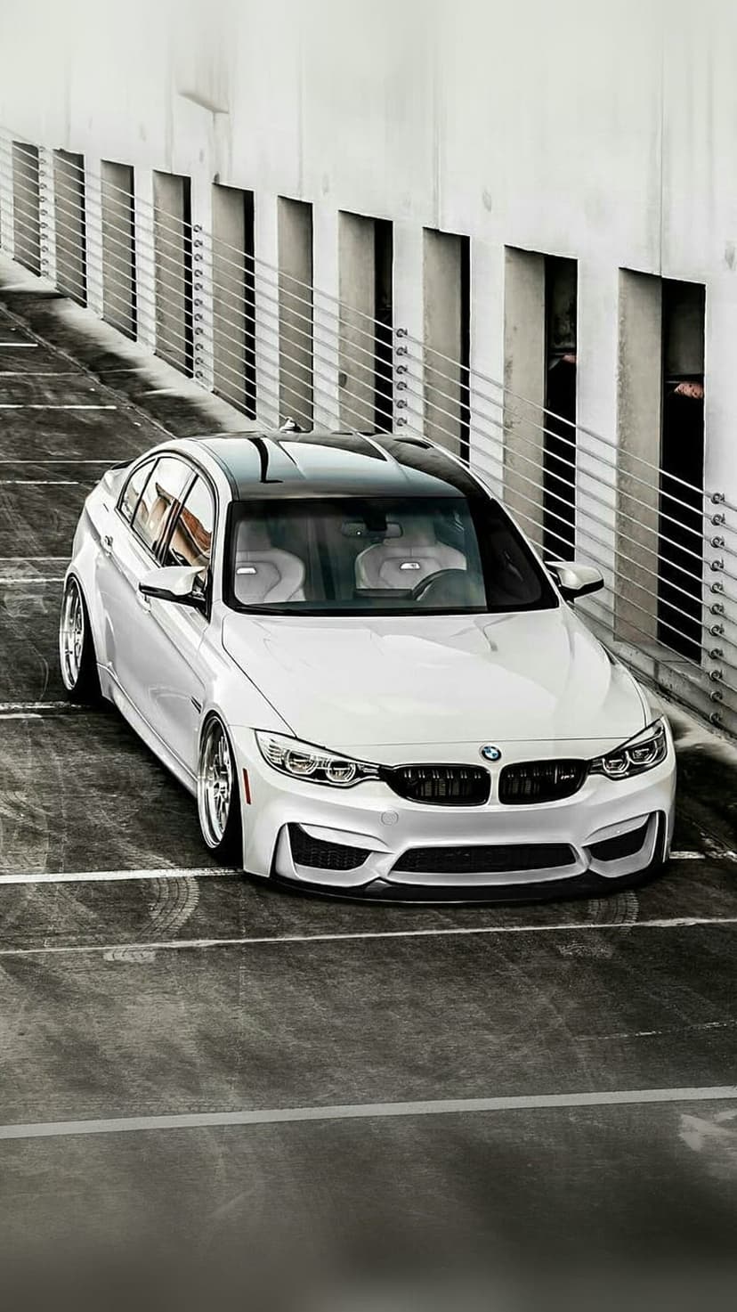 White BMW M4 parked in a modern garage featuring a black roof, custom wheels, and metallic railings at a low angle.