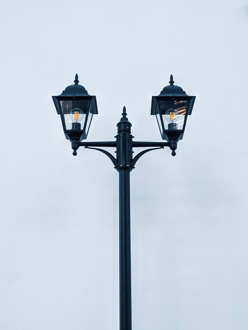 Black double lamp post with glowing bulbs centered against a pale blue sky in a vertical composition.