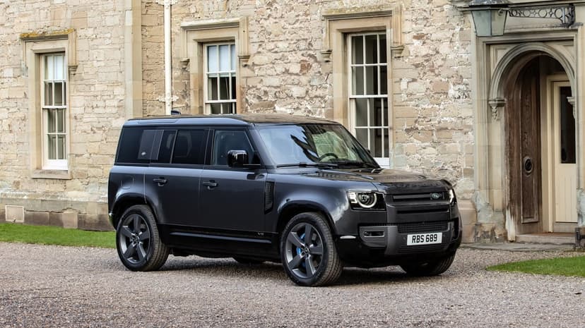Dark grey Land Rover Defender V8 parked on gravel in front of a historic stone manor house with large windows.