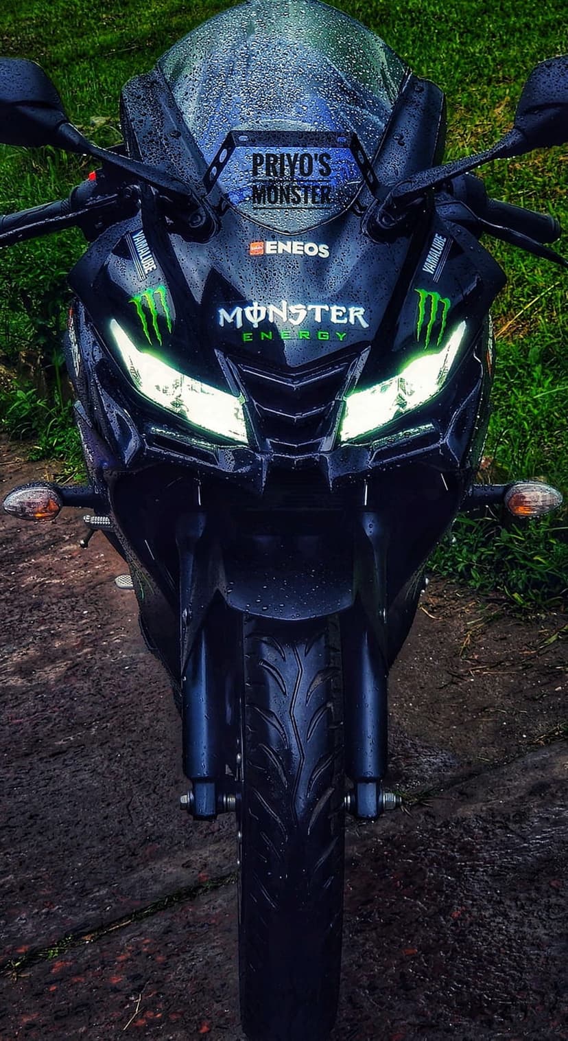 Yamaha R15 motorcycle with glowing headlights and Monster Energy decals parked on wet grass during a rainy day.