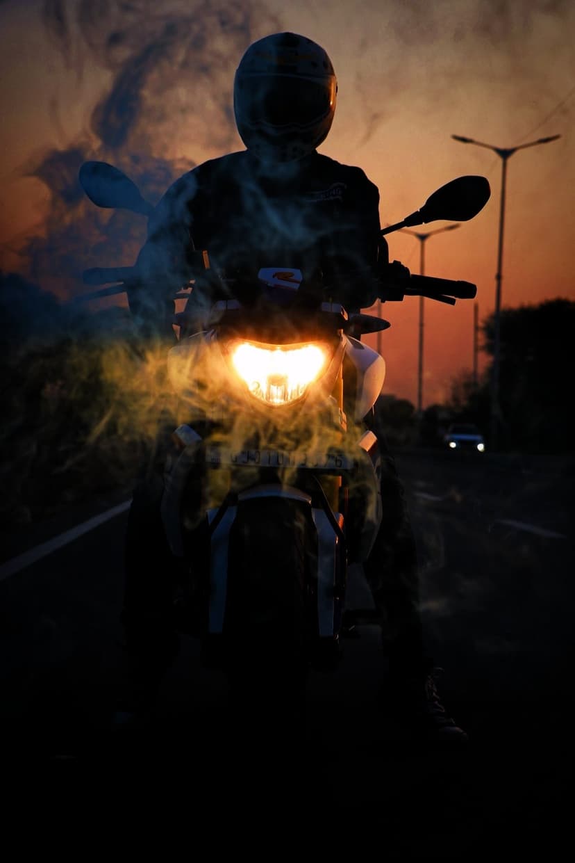 Motorcyclist silhouette with a glowing headlight amidst thick smoke against a vibrant orange and purple sunset.