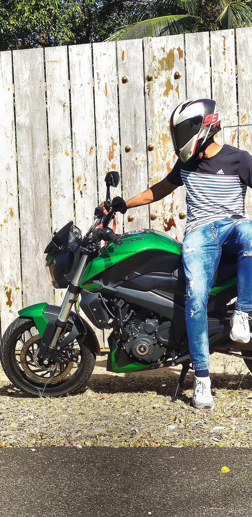 Motorcyclist in a helmet and jeans sits on a lime green bike beside a weathered wooden fence in the sun.