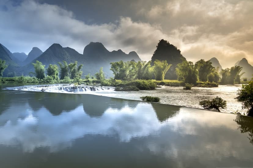 Misty limestone karst peaks tower over a tranquil river and bamboo forest with a small cascading waterfall.