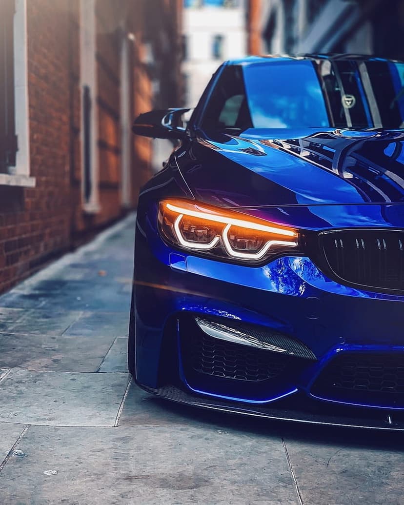 Blue BMW M4 Coupe with a large kidney grille parked on a cobblestone street beside a brick wall in a narrow alley.